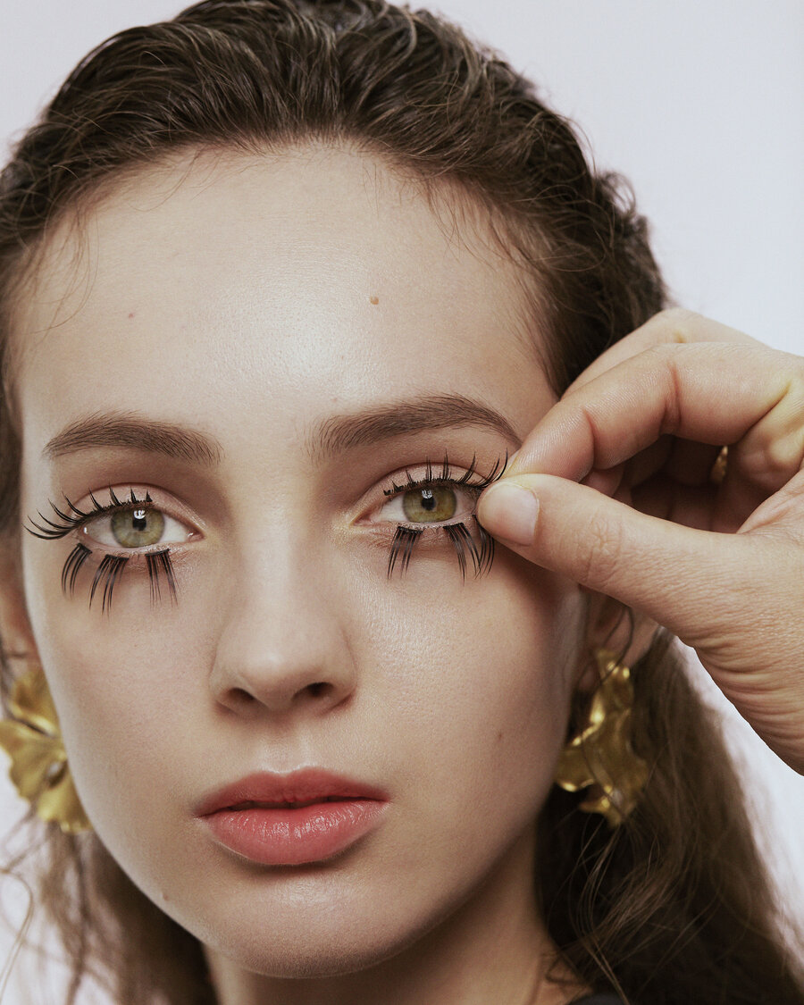 Portrait of a woman wearing gold earrings. A hand from outside the frame is applying her eyelashes