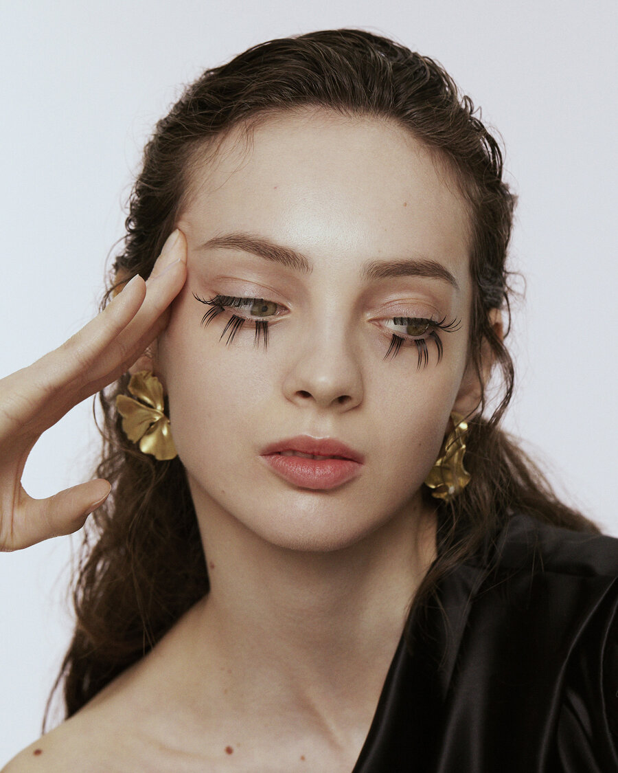 Portrait of a woman wearing gold earrings. She rests her fingers on her temple. Her eyelashes are deliberately over exaggerated
