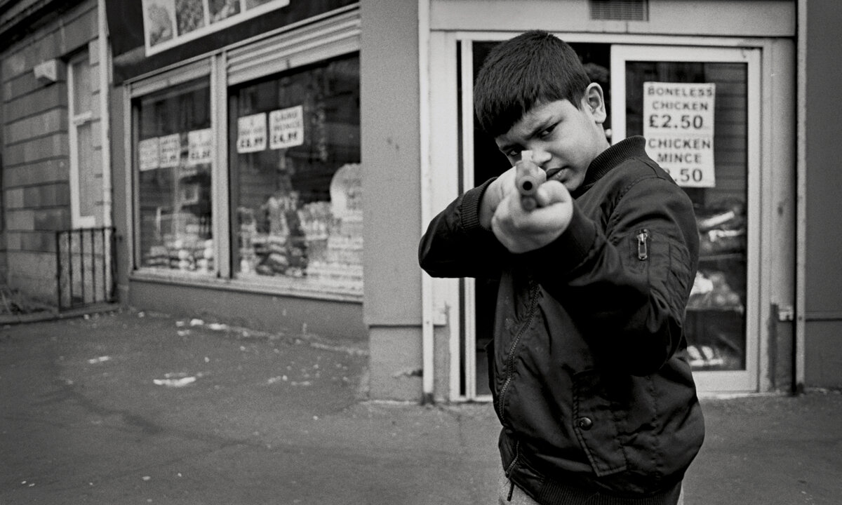 A boy stands in front of a shop pointing a shotgun at the camera. He is wearing a black jacket.